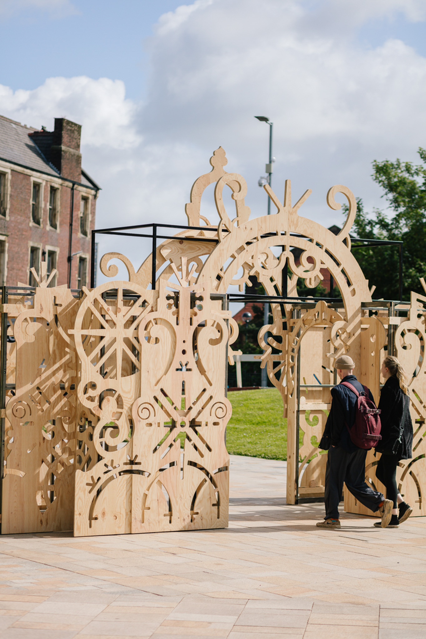 In a public square, a wooden archway made of cut out timber sheet by Tim Denton stands. Two adults walk underneath.