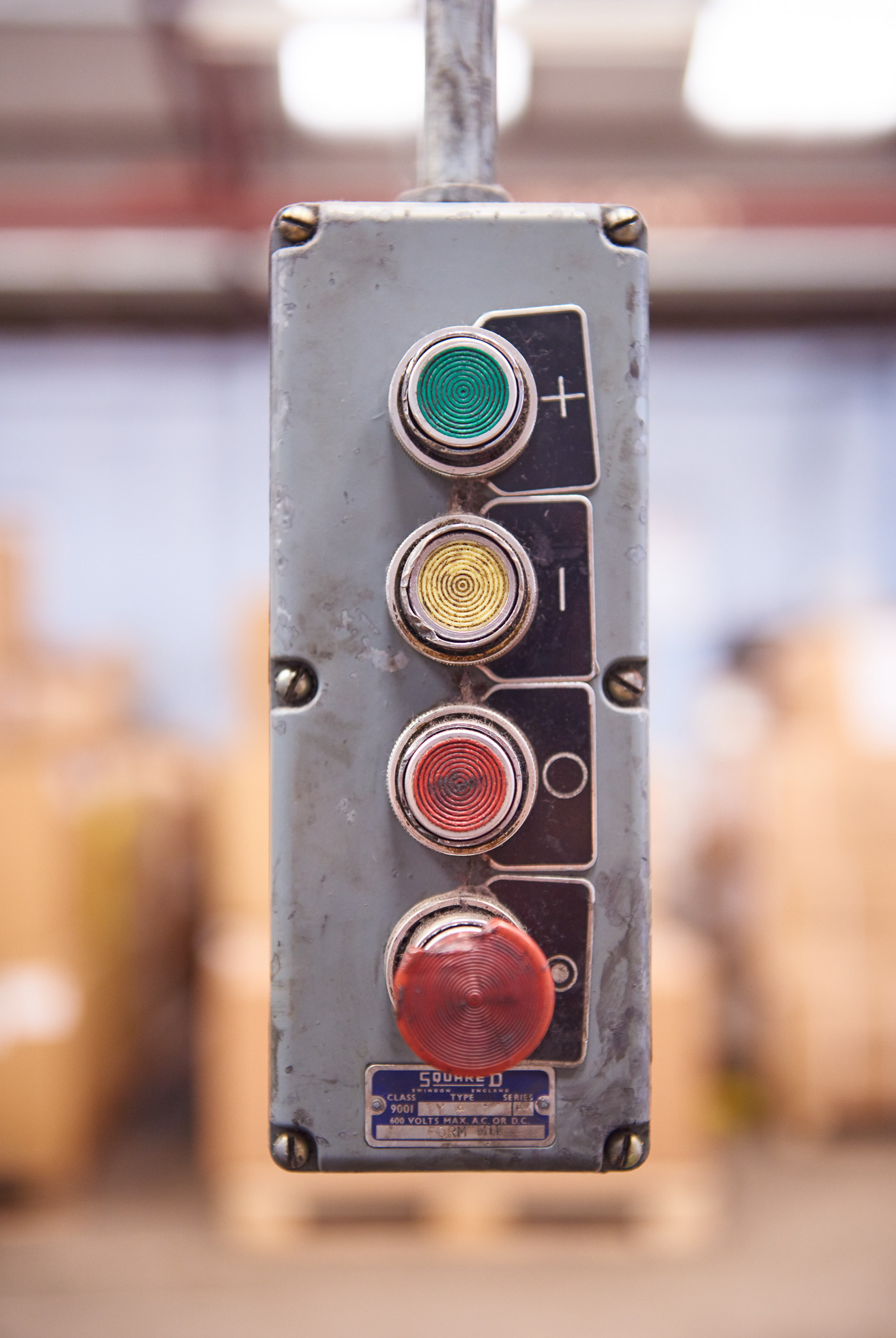 A metal control panel with four circular signal lights: green at the top, then yellow, then orange, and red at the bottom. Each has a button beside it. The background is blurred, showing an industrial setting.