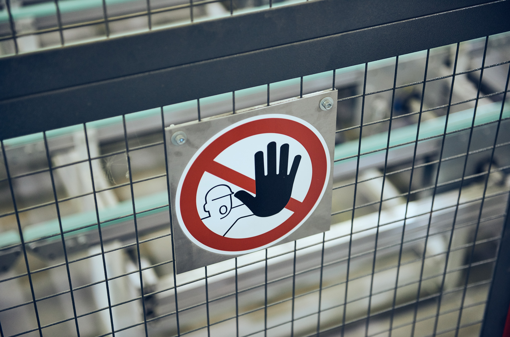 A warning sign on a wire fence shows a raised hand inside a red circle with a diagonal line, indicating no entry or do not touch. The sign also features a person wearing a safety helmet.
