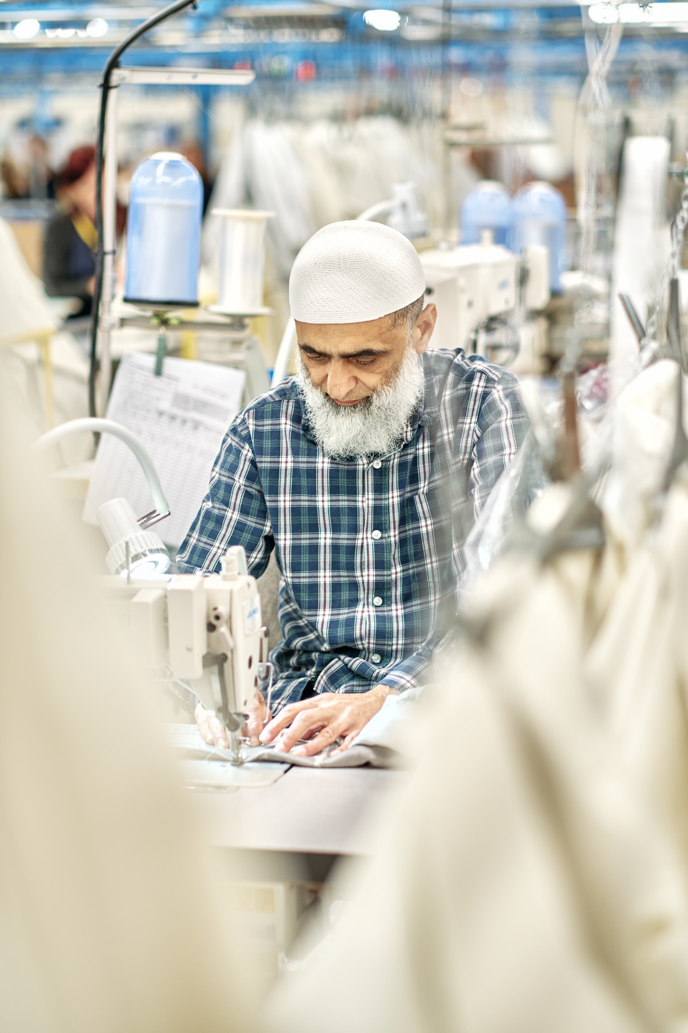 An older man with a white beard and cap, wearing a checked shirt, operates a sewing machine in a brightly lit textile factory surrounded by fabrics.