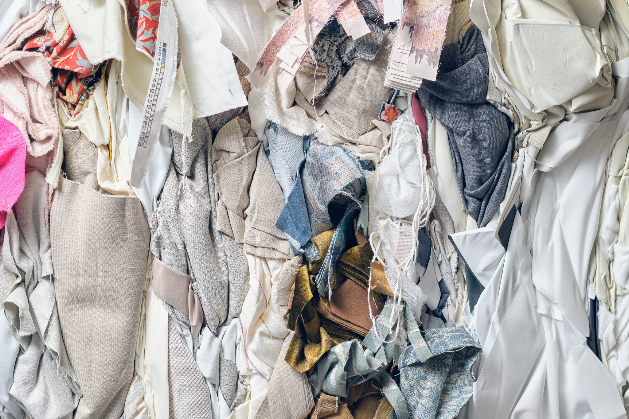 A close-up view of a pile of assorted fabric offcuts and textile remnants in various colours, textures, and patterns, including beige, white, grey, and hints of pink and gold.